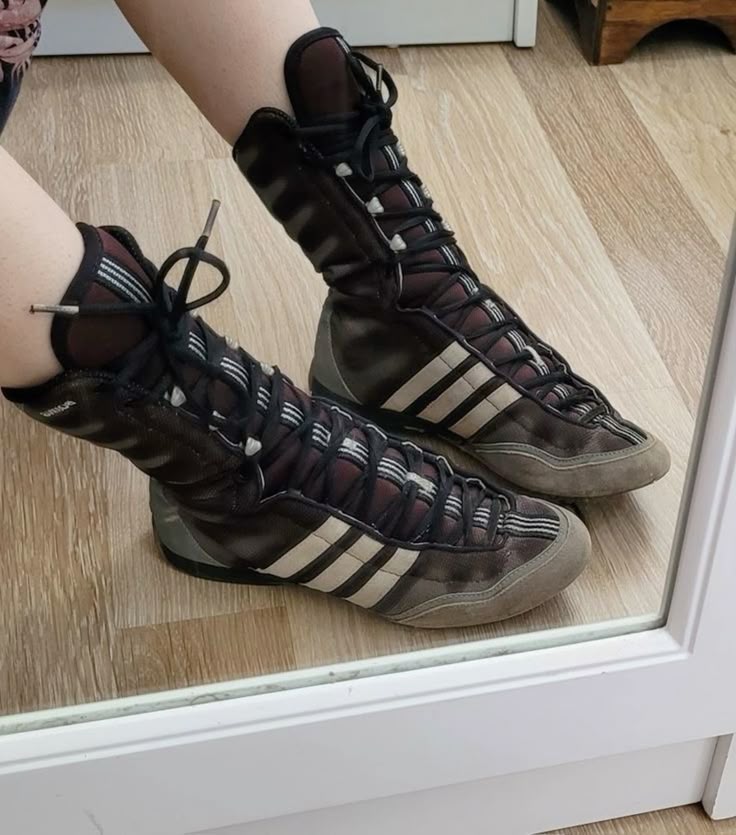 a person is wearing a pair of brown adidas wrestling shoes with white stripes. the shoes are hightops and have laces. the floor is wood. the image is reflected in a mirror.