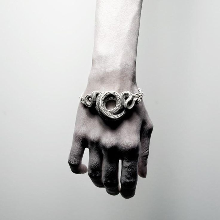 a closeup, grayscale image showcases a pale hand with a silver snake bracelet. the bracelet is coiled around the wrist, featuring intricate details of the snake's design. the hand is slightly open with fingers extending downwards. the background is a muted gray.