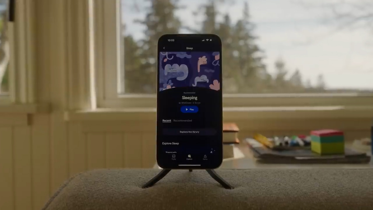 a smartphone is positioned on a stand on a piece of furniture, displaying a sleep sounds app on its screen. the background features a window with a view of trees and natural daylight illuminates the scene.