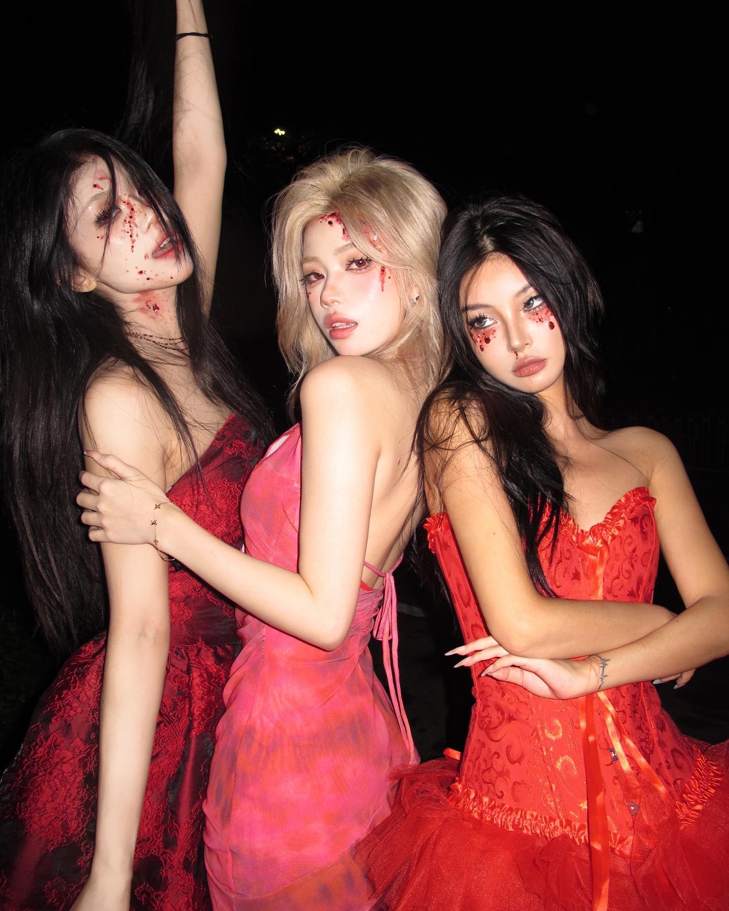 three women in red halloween costumes pose for a photo. all three have elaborate makeup looks that include fake blood. the woman on the left has long black hair and a long red dress. the woman in the middle has blonde hair and a pink and red dress. the woman on the right has black hair and a red and white dress.