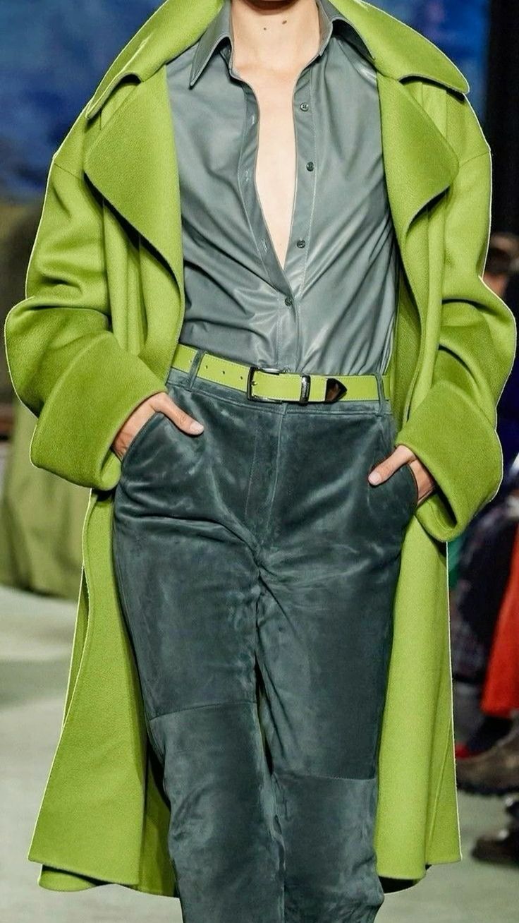 a woman is posing on a runway wearing a green coat, gray shirt, green pants and a green belt. she has a stylish and elegant look.  her outfit is a great example of current fashion trends.