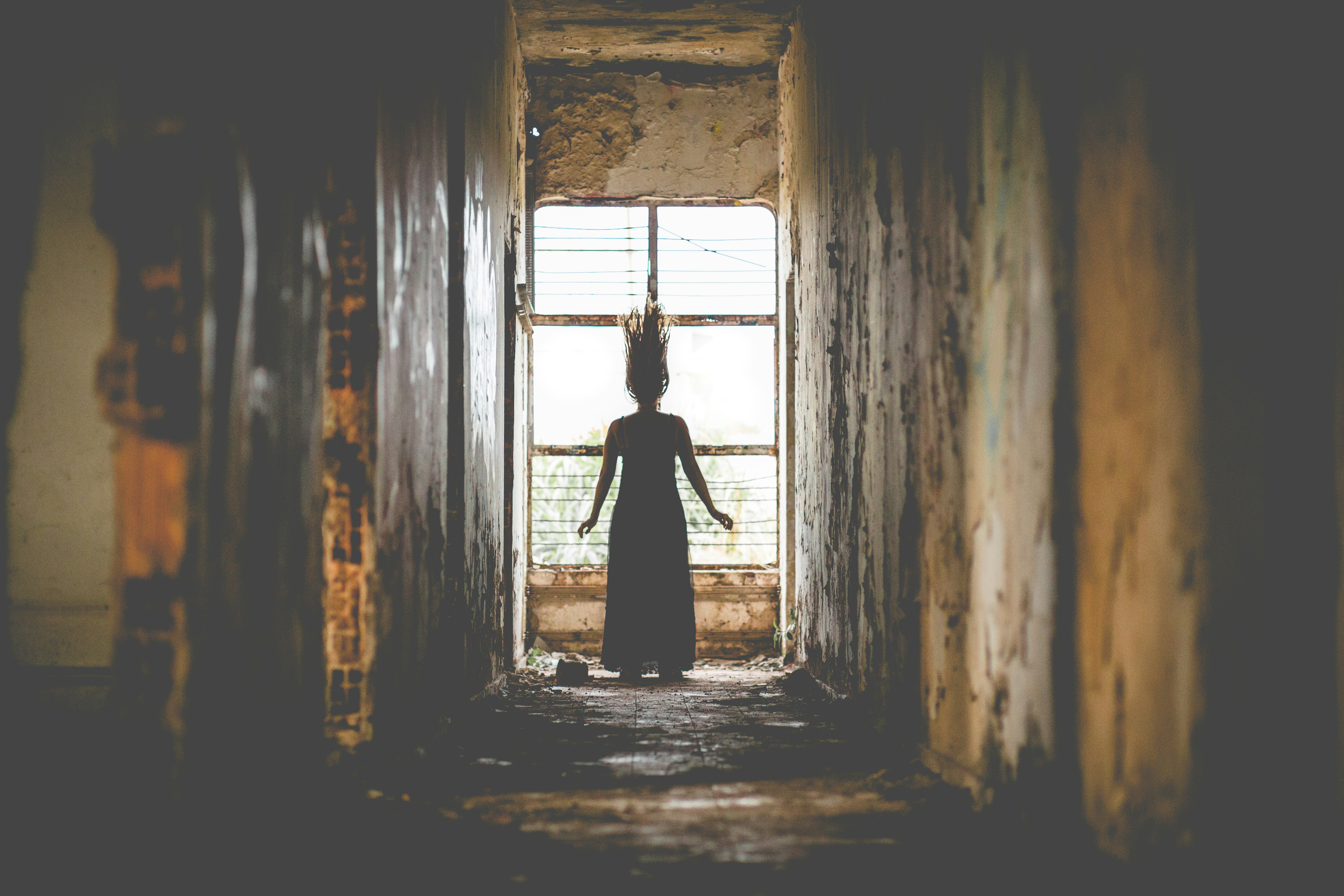 a shadowy figure of a woman in a long dress stands at the end of a dilapidated hallway. her back is to the camera, and she wears a tall, spiky crown. light streams in from a large window behind her, illuminating the dust particles in the air and creating a silhouette of her form. the walls of the hallway are crumbling and stained, hinting at a forgotten past.
