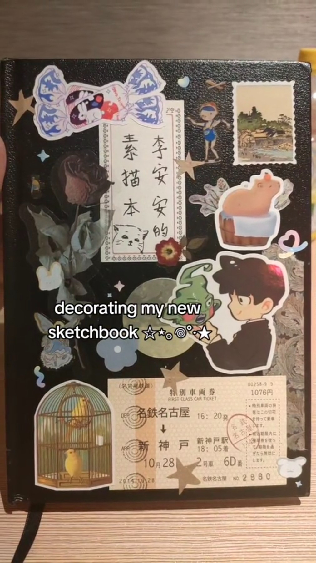 a black sketchbook decorated with stickers, including anime characters, a bird in a cage, and a train ticket.