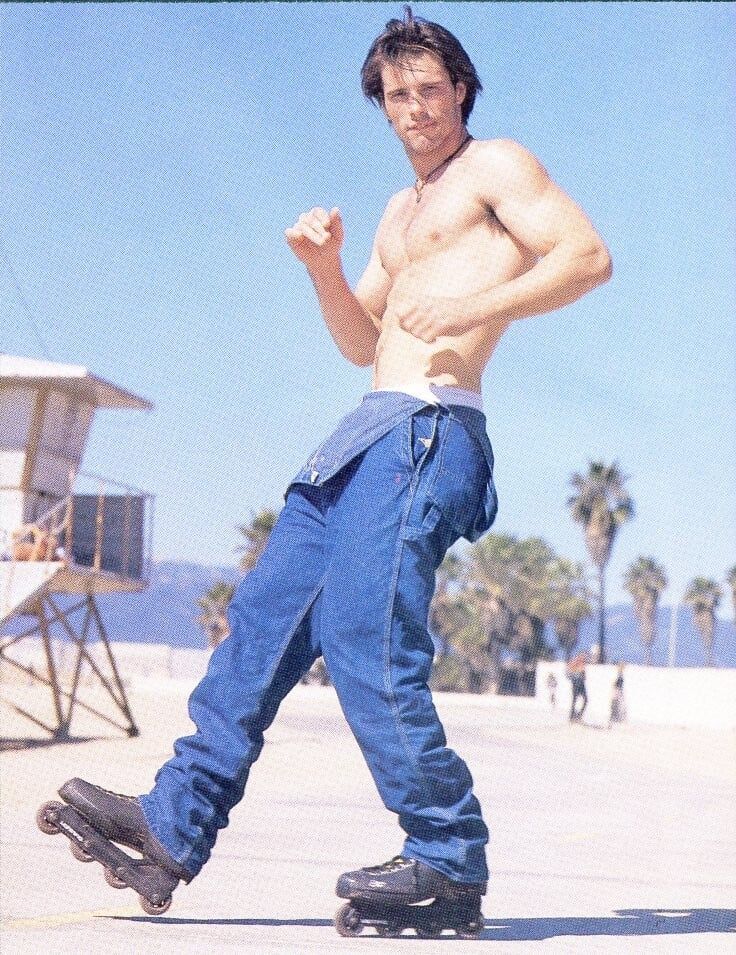 a shirtless man wearing jeans and rollerblades poses near a lifeguard station on a beach with palm trees in the background. he is looking at the camera with his fists clenched.