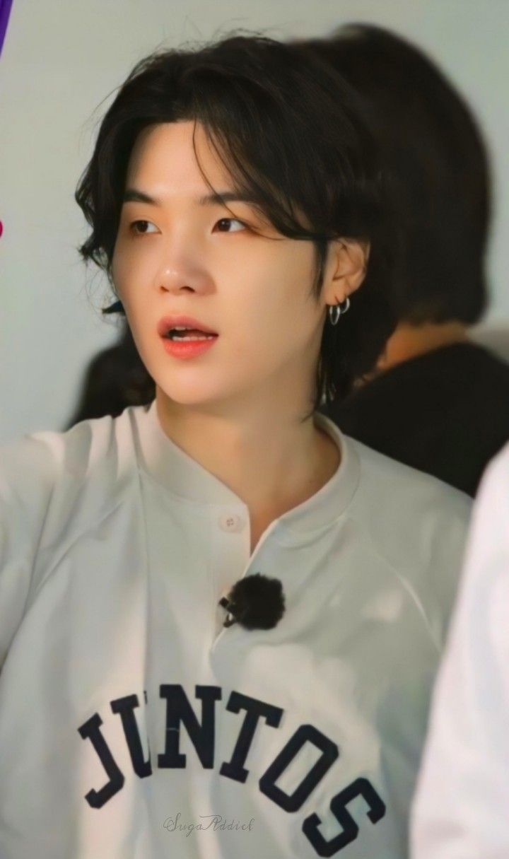 a medium shot of bts member suga, also known as min yoongi, wearing a white shirt with the word "juntos" printed on it. he has black hair styled with waves, earrings, and a small fluffy microphone clipped to his shirt. he is looking off to the side. the background is blurred. a watermark reading "suga addict" is visible on the lower part of his shirt.