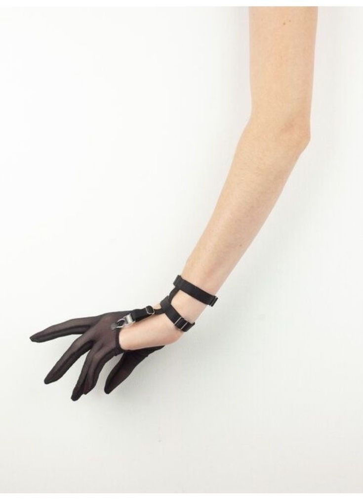 a fairskinned arm is positioned against a plain white background. the arm is adorned with a sheer black glove that covers the hand. the glove is held in place by a series of black straps with buckles that wrap around the wrist and hand. the fingers of the hand are slightly curled.