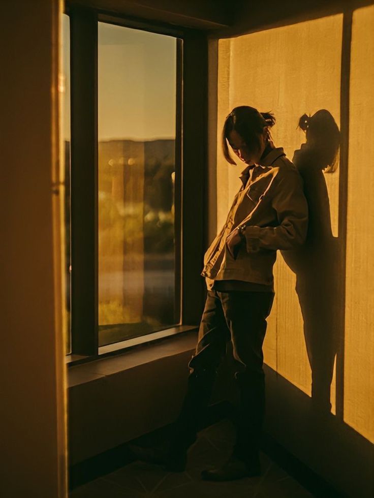 a young man is captured in a sundrenched room, illuminated by the warm, golden light of the setting sun. he leans against a wall beside a large window, his figure partially silhouetted against the bright backdrop. he's dressed in a stylish, casual outfit  a brown overshirt and dark pants. his head is bowed, and his posture suggests a contemplative mood.  the overall atmosphere is one of tranquility and quiet reflection.