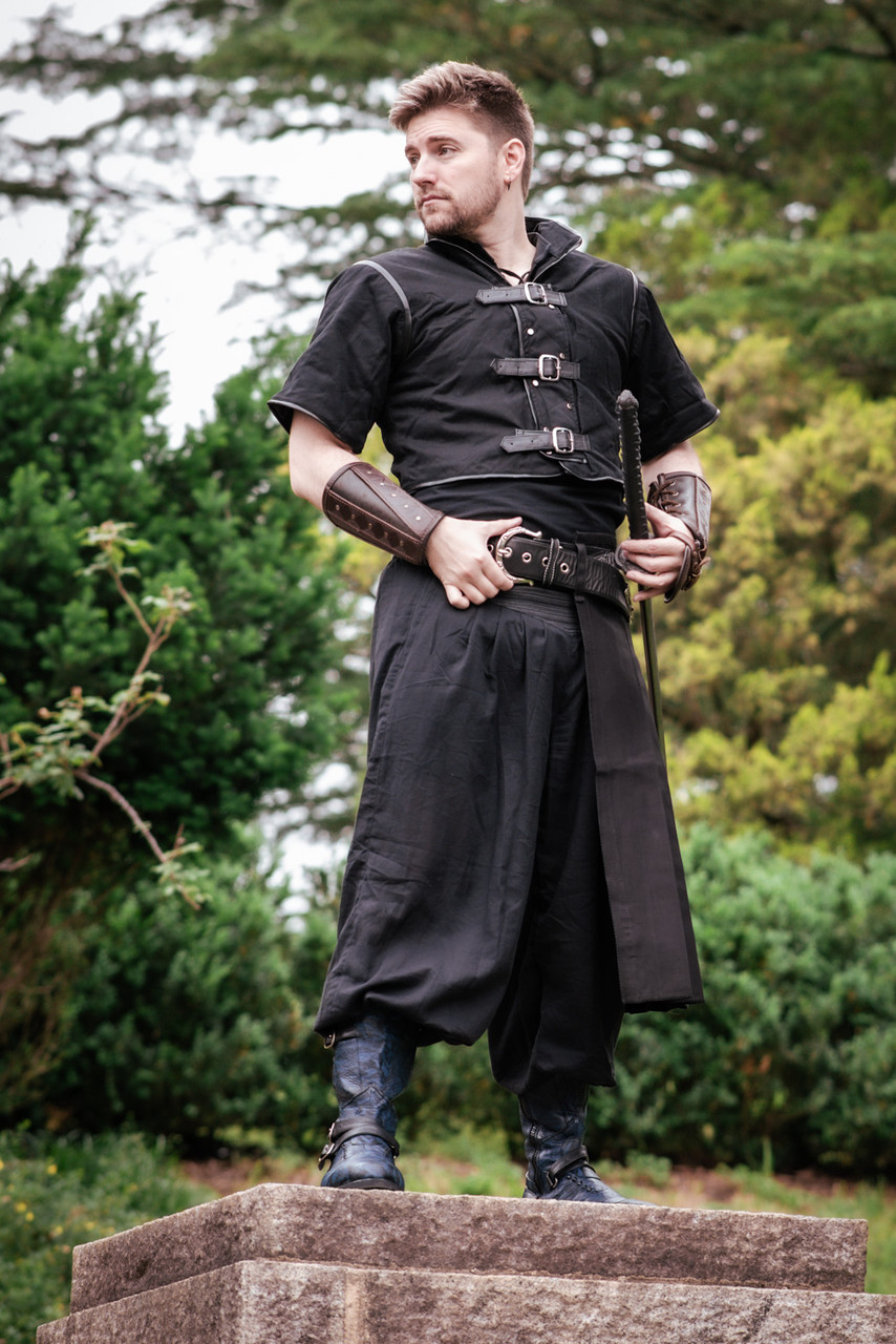 a man dressed in a medievalstyle warrior costume stands confidently on a stone platform in a park setting. he is wearing a black tunic with leather accents and buckles, loose black trousers, and blue boots. he has leather armor on his forearms and a sword in his hand. he is looking off to the side with a determined expression. the background features lush greenery, with trees and bushes providing a natural backdrop.