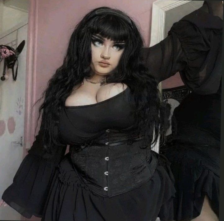 a fullfigure woman with long black hair and bangs, dressed in a black goth outfit with a corset, poses in a room with pink walls. the outfit includes a black dress and a corset, and she is wearing a necklace. decorative items are visible in the background, including a mirror.