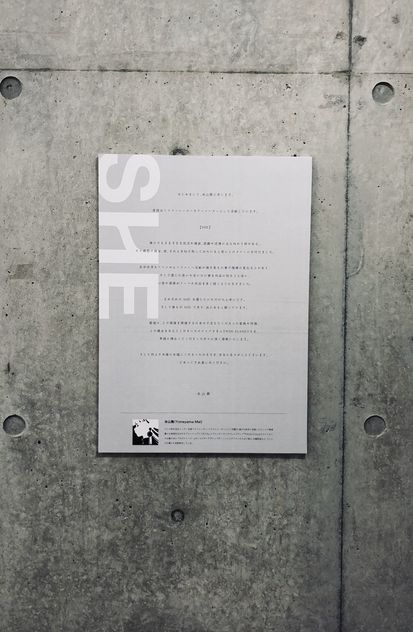 a white poster with japanese text hangs on a textured concrete wall. the poster has a black and white photo at the bottom. the lighting is soft and even, likely artificial, and the overall impression is minimalist and subdued.
