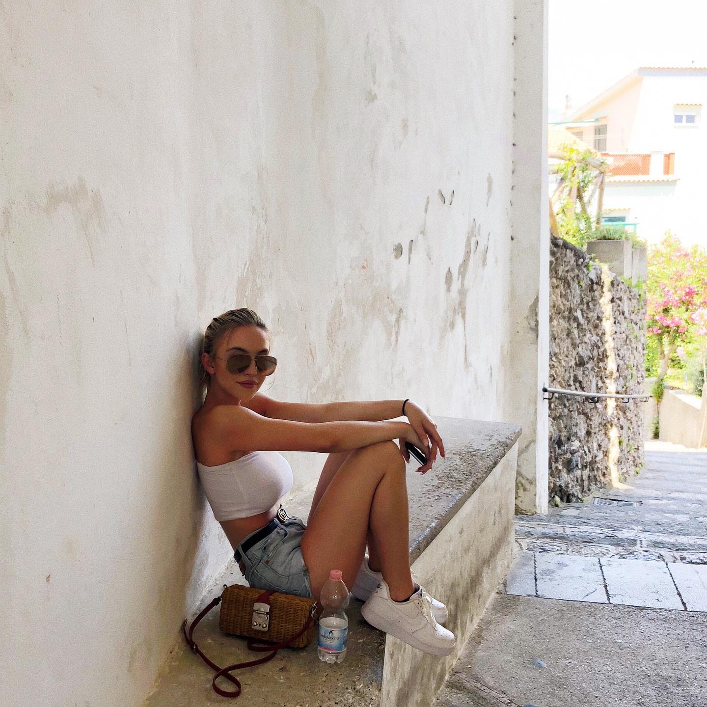 a young woman with blonde hair is sitting on a stone bench in a narrow alleyway.  she's wearing sunglasses, a white crop top, denim shorts, and white sneakers. she has a small straw handbag beside her and a water bottle next to her. the alleyway walls are white and textured; a stone wall is visible on the right, suggesting a picturesque italian setting, perhaps positano. the overall atmosphere is relaxed and casual. the image is a portraitstyle shot of the woman, capturing her from the thighs up.