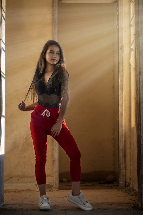 a woman poses in a doorway with light rays streaming in. she is wearing red pants, a black lace top, and white sneakers. she is looking away from the camera and holding a strand of her hair.