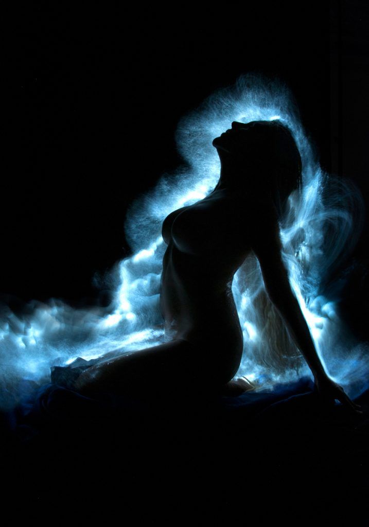 the image is a striking piece of light painting photography. a female silhouette is captured in a pose that suggests a sense of serenity or release. her body is outlined against a dark background, and her hair appears to be dynamically flowing, creating a cloudlike effect of blue light around her. the light accentuates the curves of her body, while the darkness keeps the image mysterious and evocative. the overall mood is ethereal and sensual. the technique creates a dreamlike quality, suggesting movement and energy within stillness.