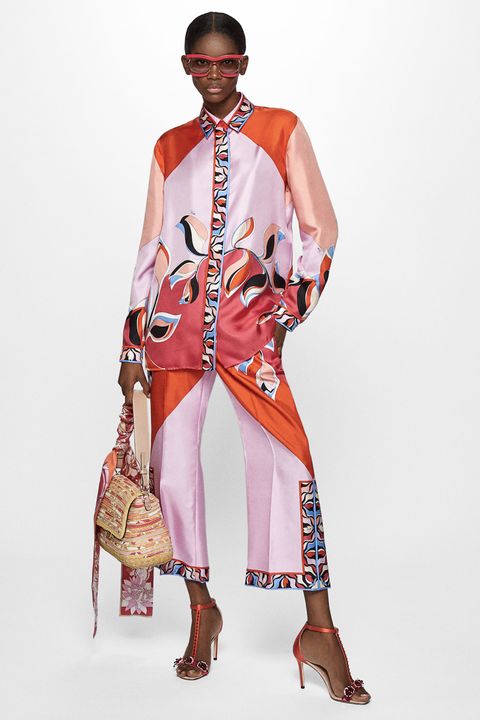 a fullbody studio shot of a stylish woman wearing a vibrant patterned silk shirt and pants set. the outfit features a color palette of pink, orange, and red with geometric and floral patterns. she's accessorized with red sunglasses, highheeled sandals, and a woven bag. the model is posing against a white background, showcasing the intricate design and details of her clothing.