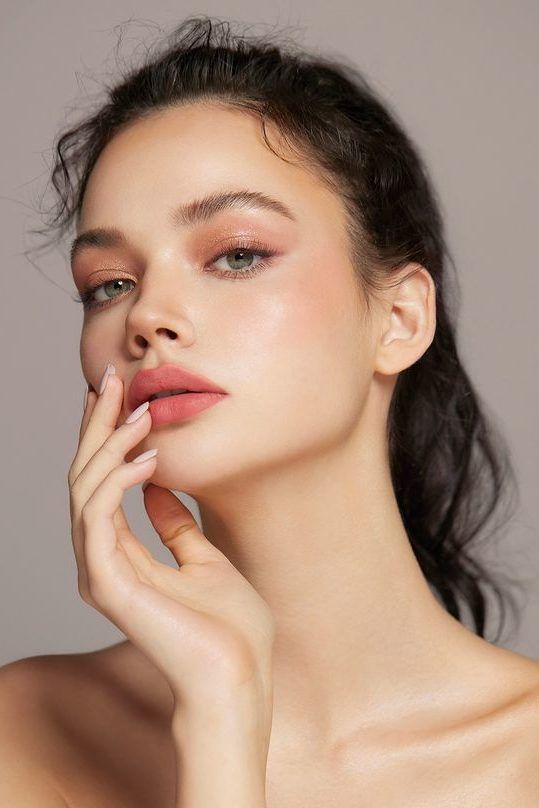 a portrait of a beautiful woman with green eyes and flawless skin. she is wearing pink lipstick and eyeshadow, and her hand is resting on her face. she has dark hair pulled back in a ponytail.