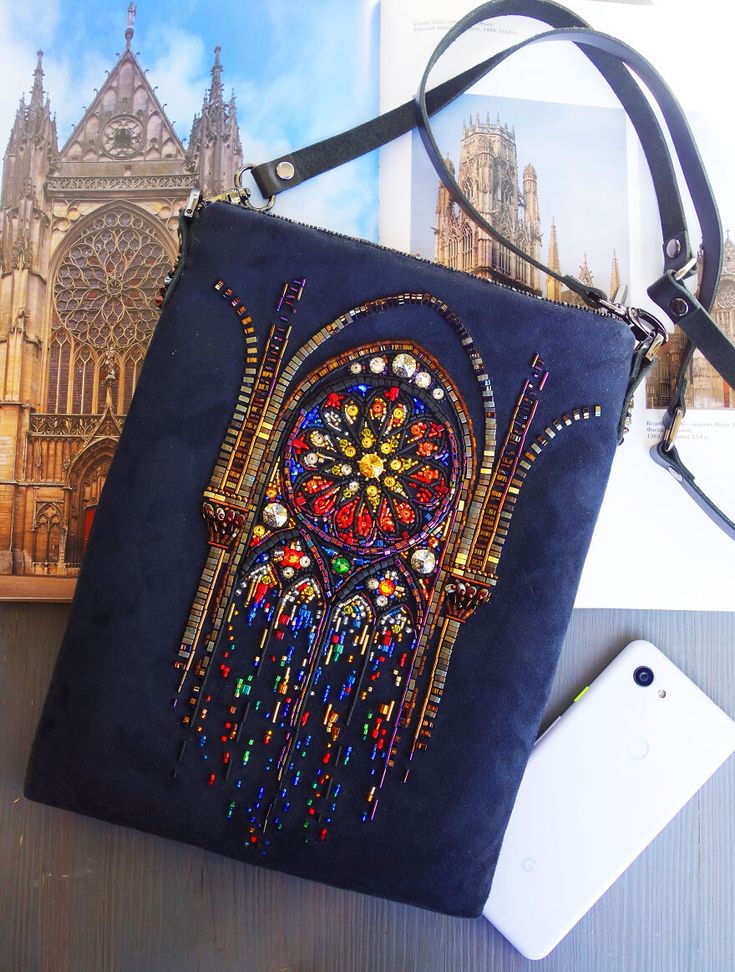 a dark blue suede bag with a stained glass window design embroidered on the front with colorful beads, rhinestones, and sequins. the bag is lying on a white surface with a phone and a magazine open to a page showing a cathedral in the background. the bag has a long black leather strap and is adorned with a unique and elegant design. it's a beautiful handmade piece with a vintage and gothic aesthetic.