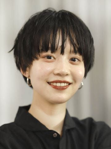 a closeup headshot of a young asian woman with short black hair and bangs. she is wearing a black collared shirt and has a natural makeup look. she is smiling and looking directly at the camera. the background is a blurry white wall.