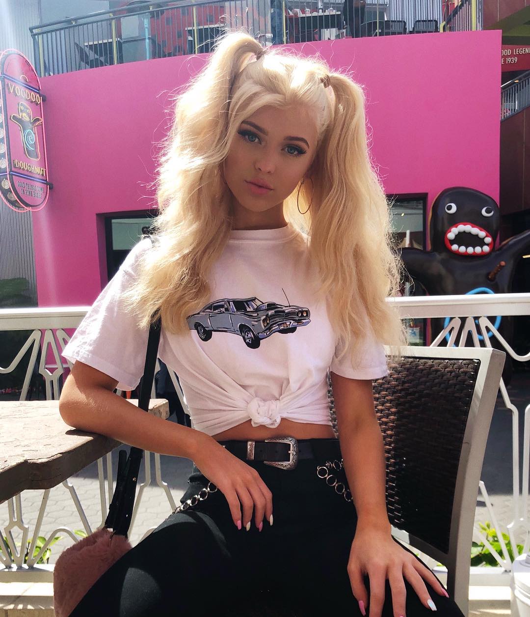 a young woman with blonde hair styled in high pigtails is seated outdoors. she's wearing a white tshirt with a car graphic, black pants with a belt and chain detail. behind her, there's a pink building, a voodoo doughnut sign, and a black statue. she is seated in a seating area with a chair and a wooden table.
