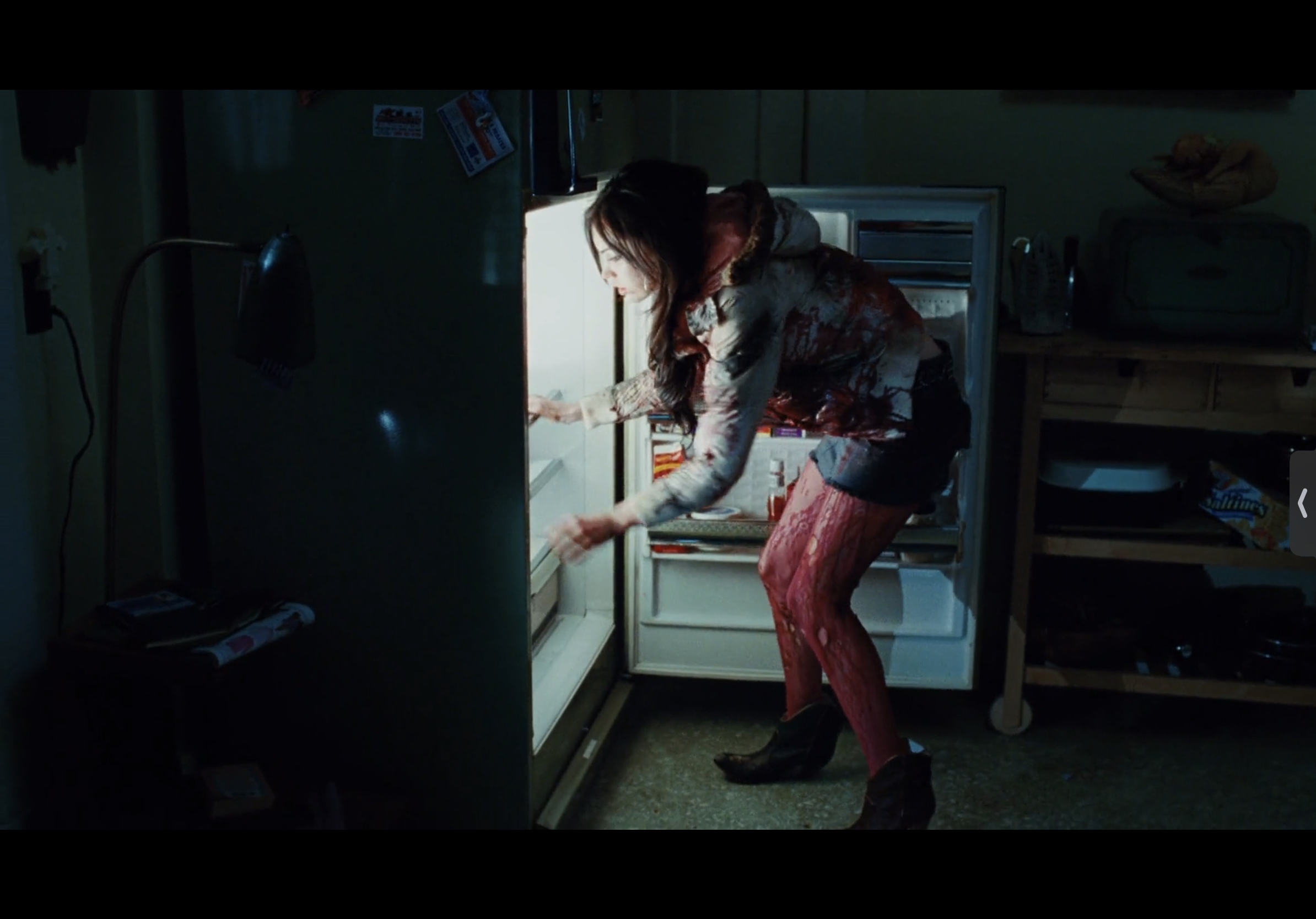 a woman with red stains all over her clothes stands with her back to the camera, reaching into an open refrigerator. the woman's clothes are stained in blood and are very messy. the refrigerator is in a dark room with only the light from the open refrigerator door.  the overall image is very dark and has a creepy/horror theme.