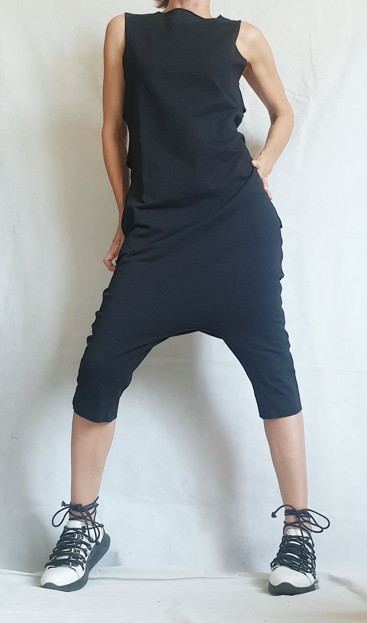 a woman stands in front of a plain white background, wearing a black sleeveless top with a pair of black harem pants. the woman is wearing a pair of black laceup white shoes.