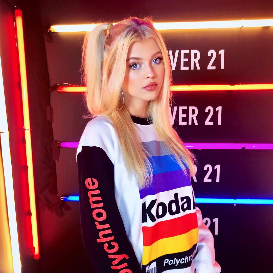 a young blonde woman with blue eyes is posing for a photo. she has her hair in high ponytails and is wearing hoop earrings. she is wearing a black and white sweatshirt with a colorful kodak polychrome design. the background is black with neon lights and the words "ver 21" repeated.