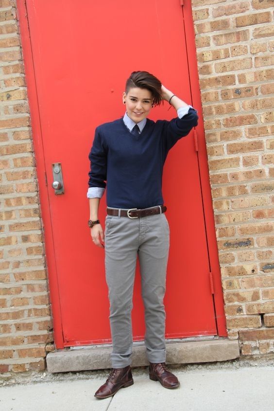 the image shows a young woman with a short, undercut hairstyle wearing a navy blue sweater over a light blue collared shirt, grey chinos, a brown leather belt and brown oxfords. she is posing in front of a red door and a brick wall. her style is androgynous, combining masculine and feminine elements. the overall impression is one of casual chic street style.