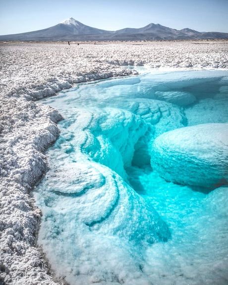 a stunning view of the salar de uyuni salt flats in bolivia. the bright turquoise water fills a crevice in the white salt, creating a surreal and beautiful contrast. the mountains in the distance add to the dramatic landscape.