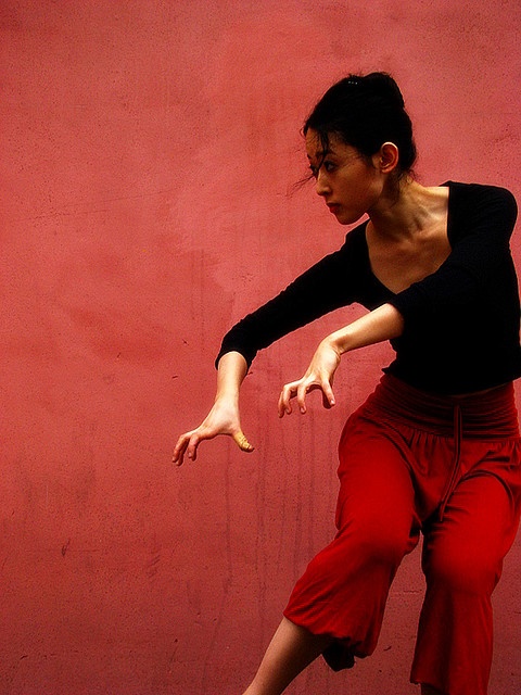 a young woman in a black shirt and red pants poses dramatically against a red wall. she has her arms outstretched and is looking to her side. her hair is pulled back and her expression is serious.