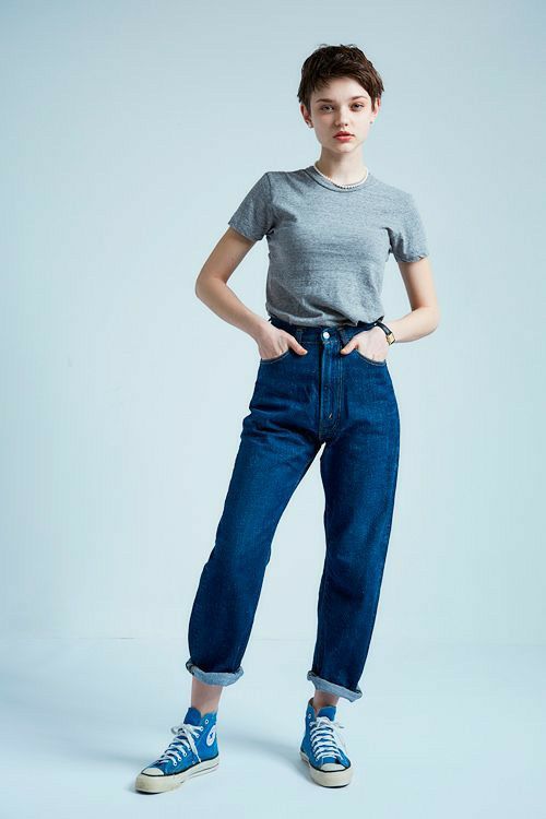 a fulllength shot of a young woman with short brown hair standing against a light blue background. she is wearing a heather grey tshirt, highwaisted dark wash blue jeans cuffed at the ankles, and light blue converse hightop sneakers.  her hands are in her pockets. she looks directly at the camera with a neutral expression. the image is welllit and clean, typical of fashion photography.