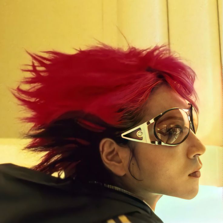 a young woman with pink hair wearing sunglasses looks away while looking serious. her hair is styled in a mohawk and she is wearing a casual outfit.