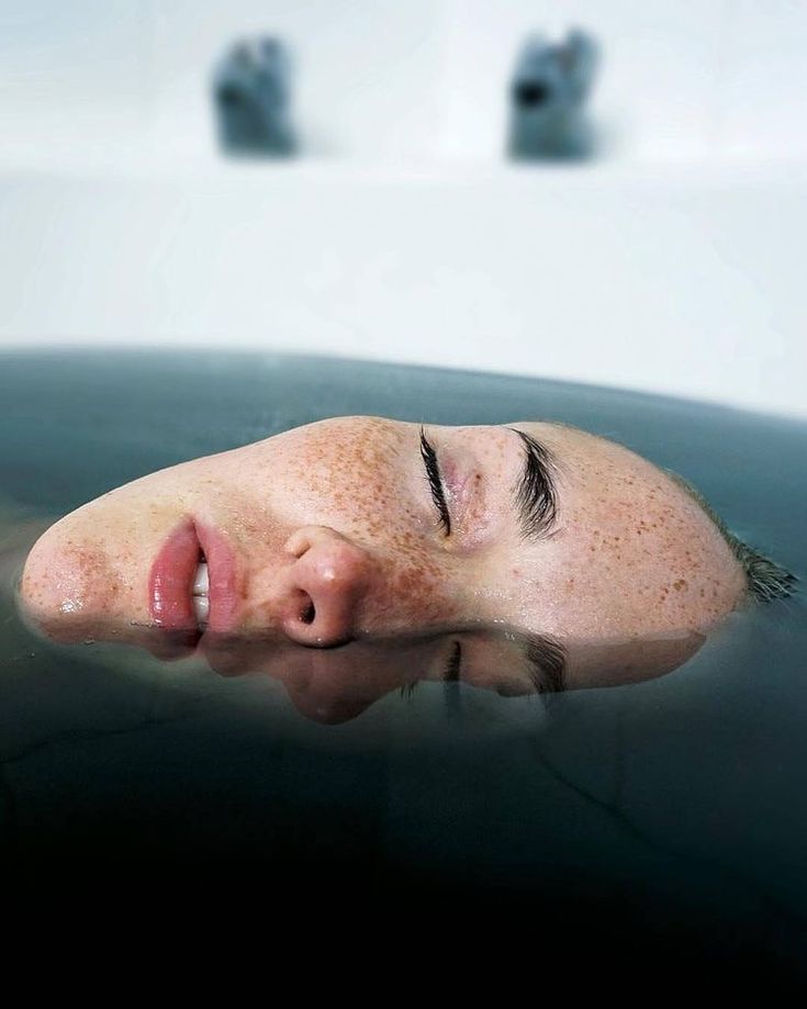 a woman's face is partially submerged in a dark, murky water. her eyes are closed, and she appears to be asleep or meditating. the water is still and calm, reflecting the light of the sun. there is a sense of peace and tranquility in the image, as if the woman is at one with the water.