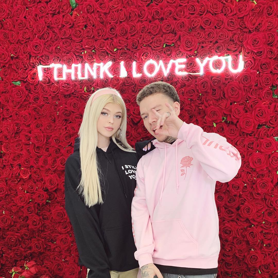 loren gray and a man stand in front of a wall of red roses with a neon sign that reads "i think i love you." loren is wearing a black hoodie with the words "i still love you" printed on it and a pink headband. the man is wearing a pink hoodie with a rose design. he is making a hand gesture with his fingers.