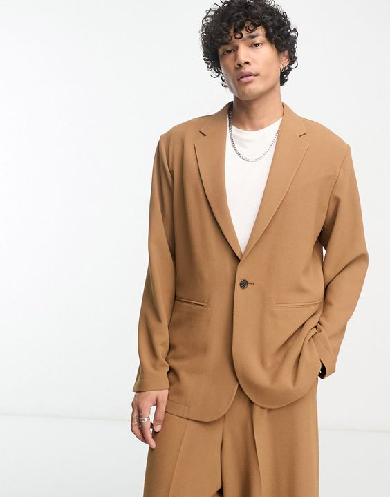 a young man is posing for the camera wearing a brown suit with a white shirt. he is looking directly at the camera with his arms at his sides. he is wearing a silver necklace. the background is white.
