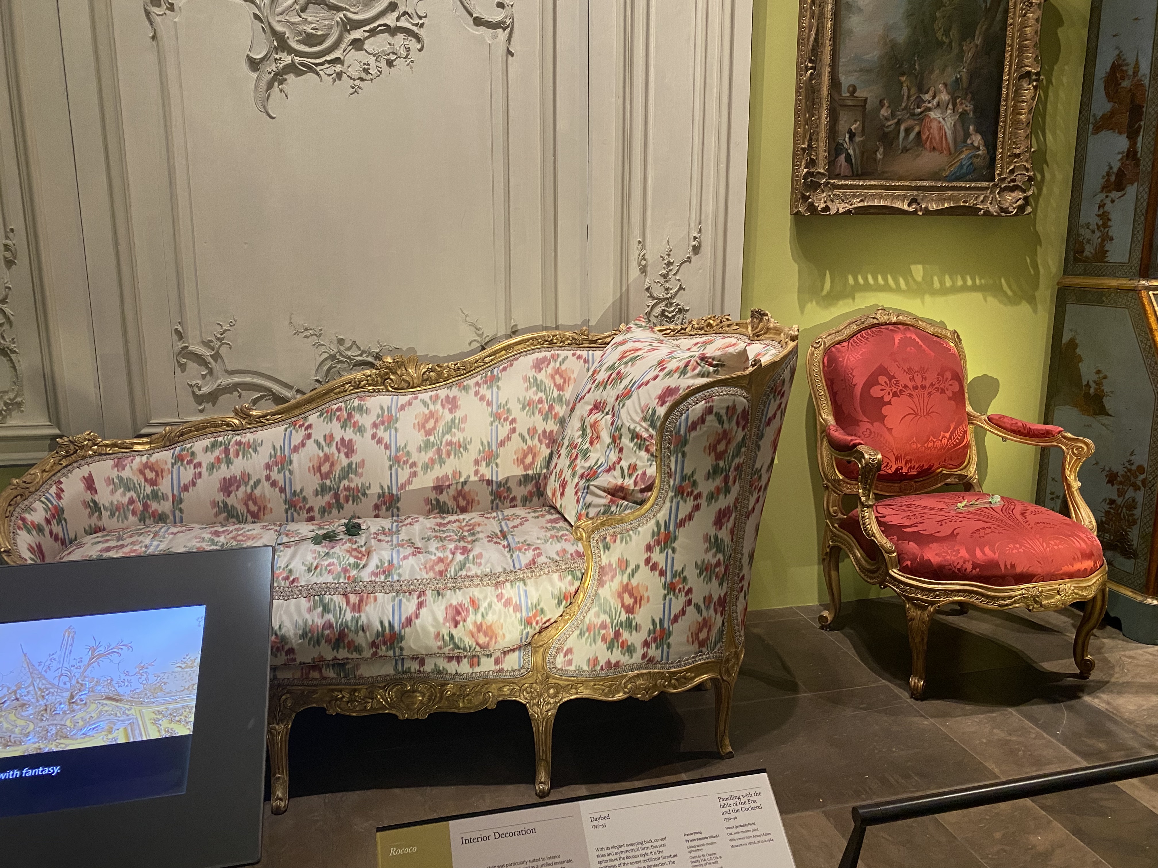 the image shows a rococostyle interior decoration exhibit in a museum setting. a floralpatterned couch with a gold frame is placed next to a red chair, also with a gold frame. the wall is decorated with intricate plasterwork and a painting hangs above the chair. a display panel is visible in front of the couch, providing information about the exhibit.
