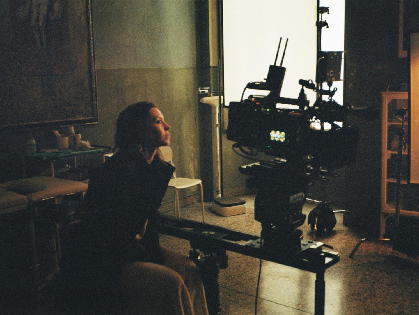 the image is a behindthescenes shot on a film set. a young woman, possibly an actress, sits pensively in a dimly lit room that appears to be a repurposed hospital or medical setting. she is dressed in dark clothing and looks thoughtful, gazing off to the side.  in the foreground is a professional film camera mounted on a dolly, ready for filming. various lighting equipment and other film set paraphernalia are visible around the room, creating a moody and atmospheric scene. the overall aesthetic is dark, gritty and cinematic, suggesting a film noir or similar genre.  a large, old painting hangs on the wall adding to the vintage feel of the space. the image has a grainy quality, typical of film photography.