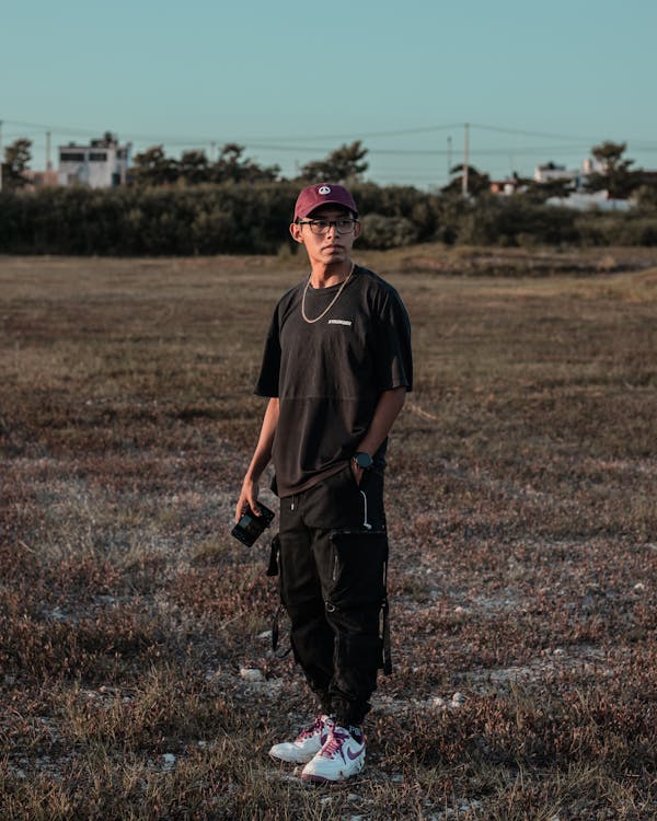 a young man stands in a field, looking away from the camera. he is wearing a burgundy cap, a black tshirt, black cargo pants, and white nike sneakers with purple accents. he has a gold necklace on and is holding a small black rectangular object in his hand. the setting appears to be a rural area in the late afternoon or early evening, as the sky is a light blue and the lighting suggests the sun is setting. the overall mood is calm and contemplative.