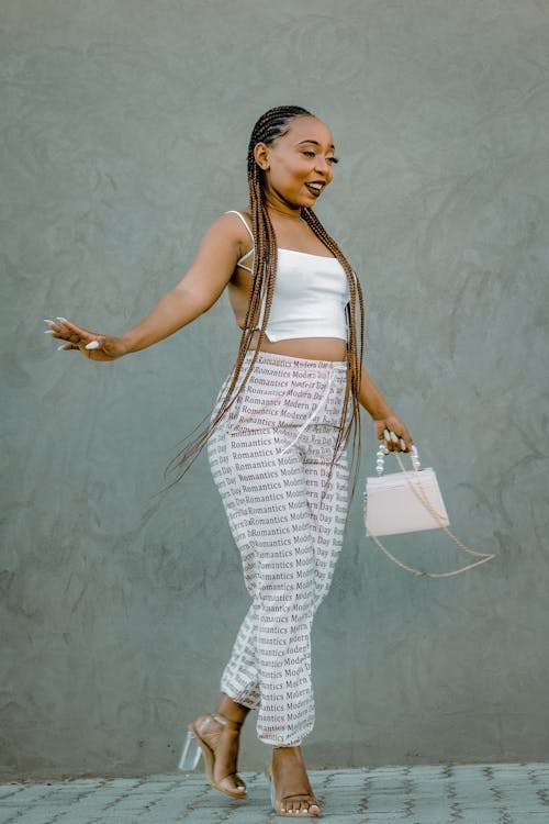 young, stylish african woman with long brown braids is walking confidently towards the right side of the image. she's wearing a white crop top,  lightcolored pants with a repeated text print, clear highheeled sandals and a light pink handbag. she's posing casually against a textured grey wall outdoors in bright daylight.  the overall impression is one of summery, casual chic style and confident femininity.