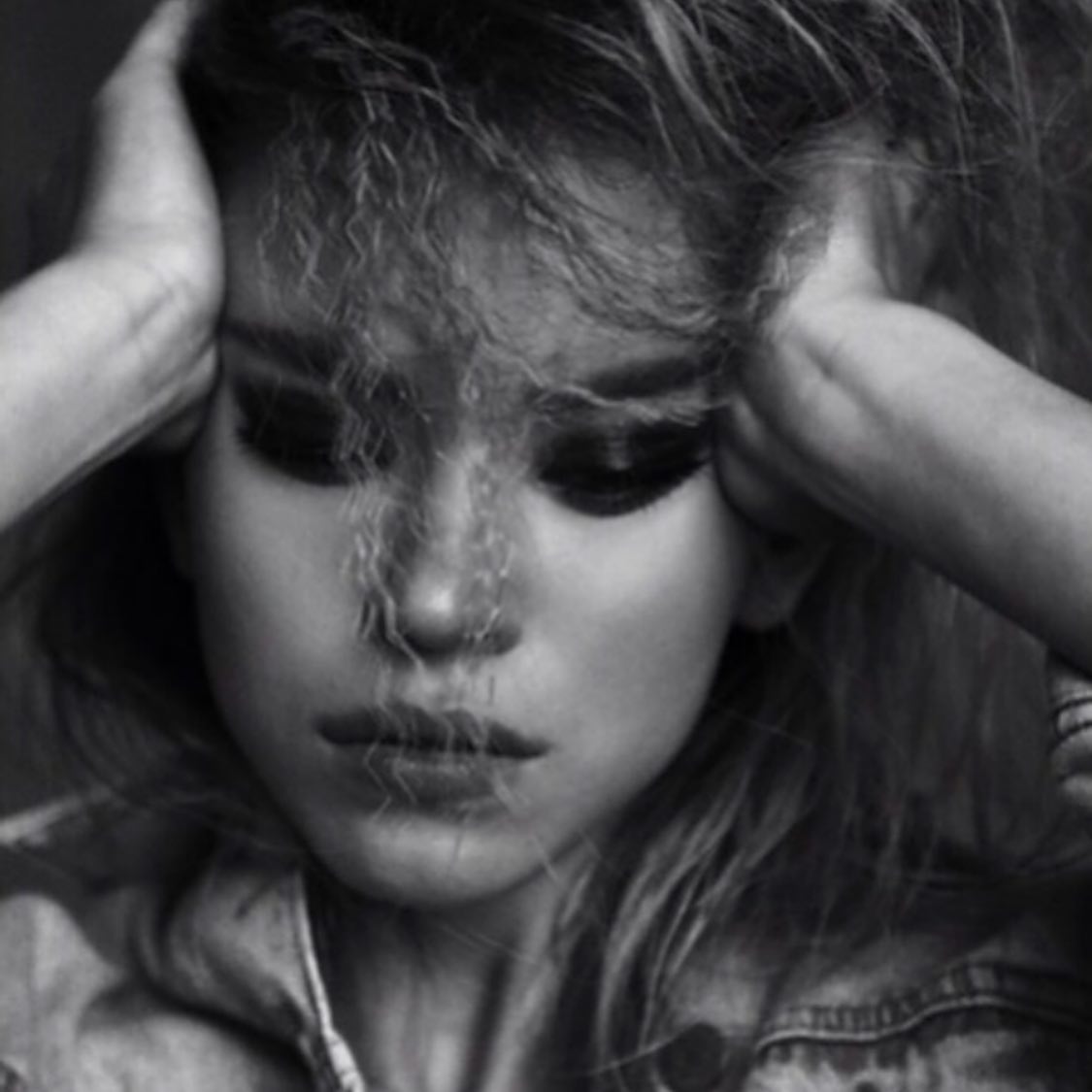 the image is a closeup, black and white portrait of a young woman with long, messy blonde hair.  she is holding her head with both hands, her expression is pensive and somewhat melancholic. her makeup is noticeable, especially the dark eyeshadow. she's wearing a denim jacket. the overall mood is moody and dramatic, with a strong focus on the woman's emotional state. the image has an artistic and slightly grunge aesthetic.