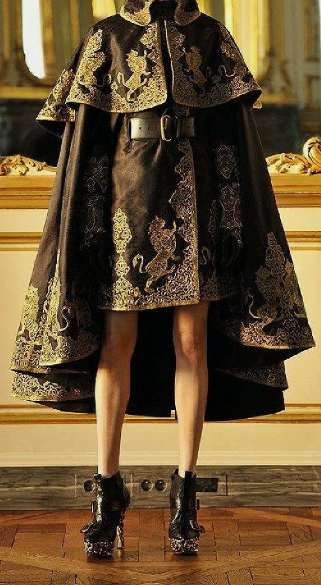 a woman in a black cape with gold embroidery stands on a wood floor. the cape is a flowing garment with a high collar and long sleeves that are tied at the wrist. it is decorated with intricate gold embroidery. the woman's dress is black and is covered in a gold design that matches the cape. she is wearing high black boots and a belt with a gold buckle. she looks elegant and glamorous in her royal attire.
