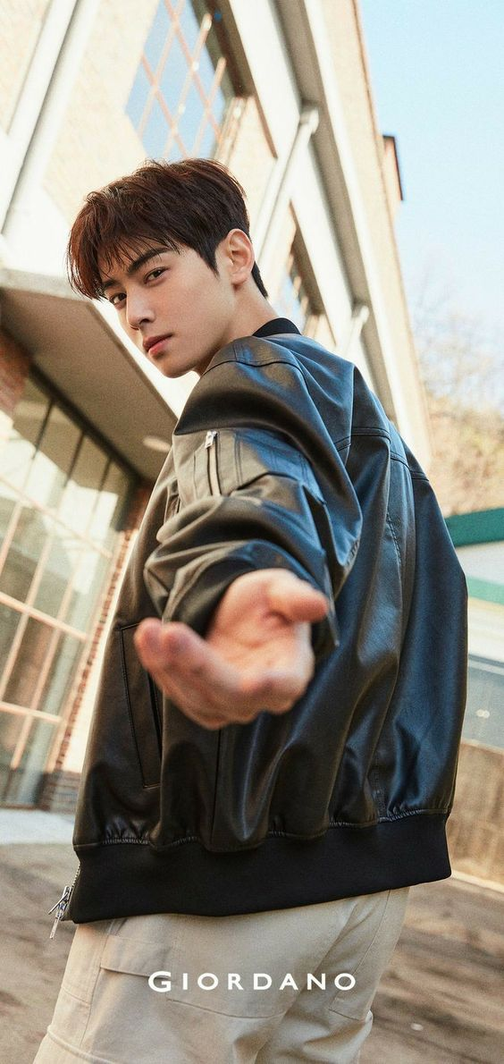 a young man, likely a model, poses in front of a brick building wearing a black leather jacket, white pants, and a chain. he extends his hand towards the viewer in a welcoming gesture. the low angle shot accentuates his height and the giordano logo is prominently displayed at the bottom of the image, suggesting this is an advertisement for the clothing brand. the sunny outdoor setting and natural lighting create a bright and inviting mood.