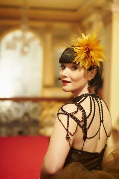 a woman is posing in a studio setting with a red carpet backdrop. she is wearing a vintageinspired flapper dress with a beaded embellishment on the back. she is also wearing a feathered hat with a yellow flower on the top. she has on red lipstick and her hair is styled in a bob with bangs.