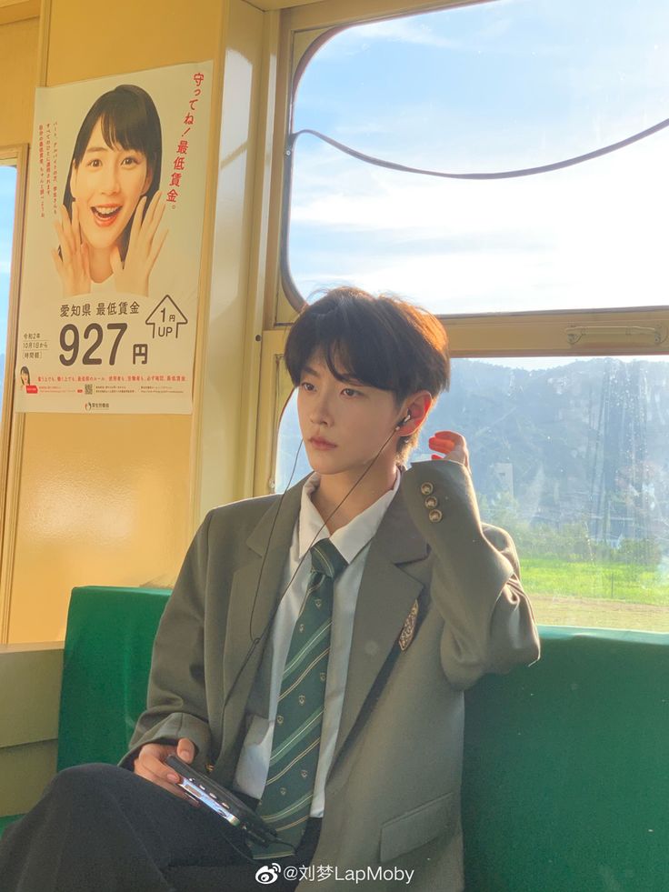 a young asian man is sitting on what appears to be a japanese train. he's wearing a stylish green suit jacket and a striped tie. he's also wearing earphones and appears to be listening to music. the train windows offer a glimpse of a scenic landscape outside. the lighting is natural and bright, giving the photo a warm and inviting feeling.