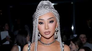 a closeup portrait of a woman with long, intricate silver box braids styled in an updo.  she's wearing  makeup with a focus on her eyes and lips.  she also has on a silver necklace and earrings and is dressed in a partially visible black and white patterned top. the background is blurry, suggesting she's at an event or party. the overall impression is one of elegance and glamour.