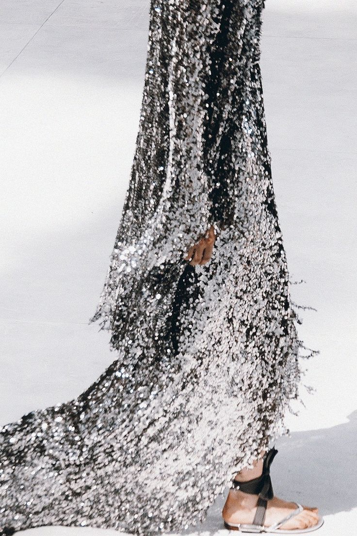 a fashion model walks the runway in a stunning silver sequin dress with a long train. the dress is extremely sparkly and catches the light beautifully. she is wearing sandals and her hair is styled in a simple way to let the dress take center stage. this is a highfashion look that is perfect for a special occasion.