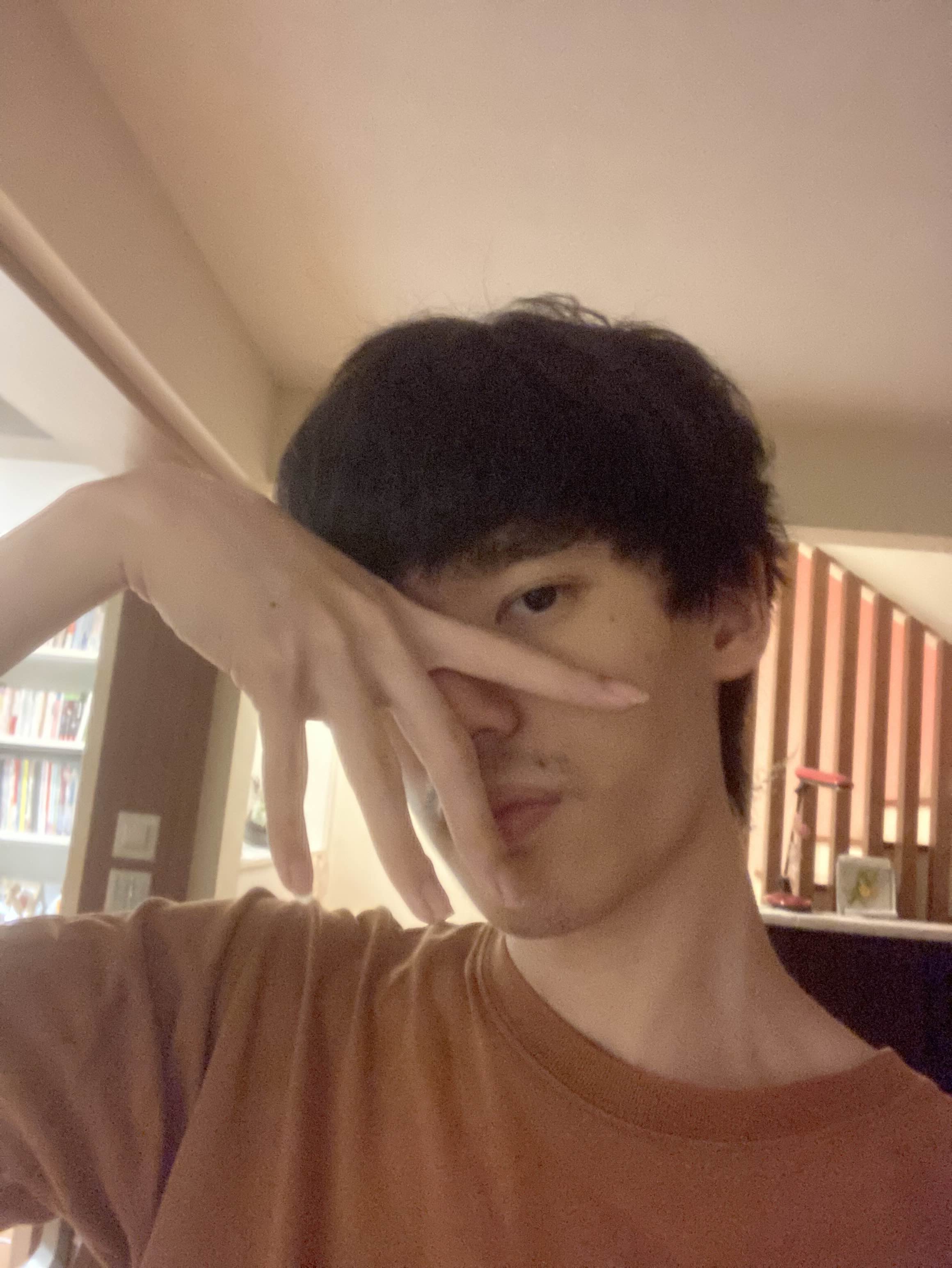 a closeup selfie of a young asian man with short dark hair. he is wearing a brown tshirt and making a playful hand gesture, extending his fingers across his face in front of one eye. the setting appears to be indoors, likely his home, with warm artificial lighting.  a bookcase is visible in the background to the left.
