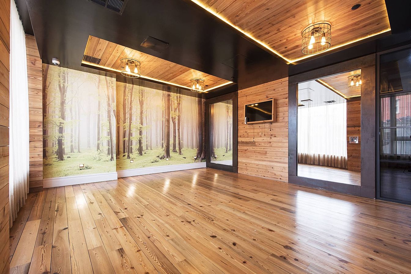 a spacious room with wooden floors, a woodpaneled ceiling, and a large mirror on one wall. the room is decorated with a woodpaneled wall, a wallpaper depicting a forest, and a large window. there are also several lamps hanging from the ceiling. the room is bright and airy, with a modern and contemporary feel.