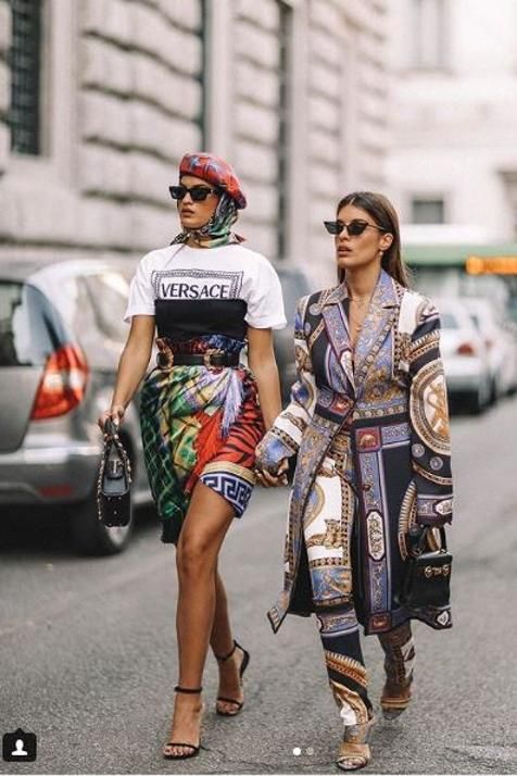 two fashionable women are walking down a city street. one is wearing a versace tshirt, a colorful patterned skirt, and black heels, while the other is wearing a long, patterned jacket and pants, along with heels.  they both carry small purses. they are both wearing sunglasses. the image is vibrant and stylish, showcasing a trendy street style look.