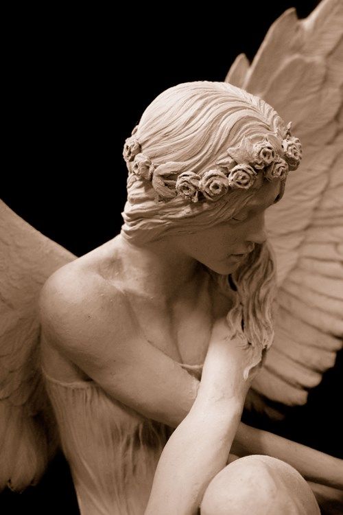 the image showcases a beautifully sculpted bust of a female angel with her head bowed and her wings gracefully curved behind her. crafted likely from clay, the sculpture is monochrome, emphasizing the play of light and shadow across the smooth, detailed surfaces. the angel wears a delicate crown of flowers and her hair flows down her shoulders, partially obscuring her face. her contemplative pose and the soft lighting contribute to the overall serene and melancholic mood of the piece.