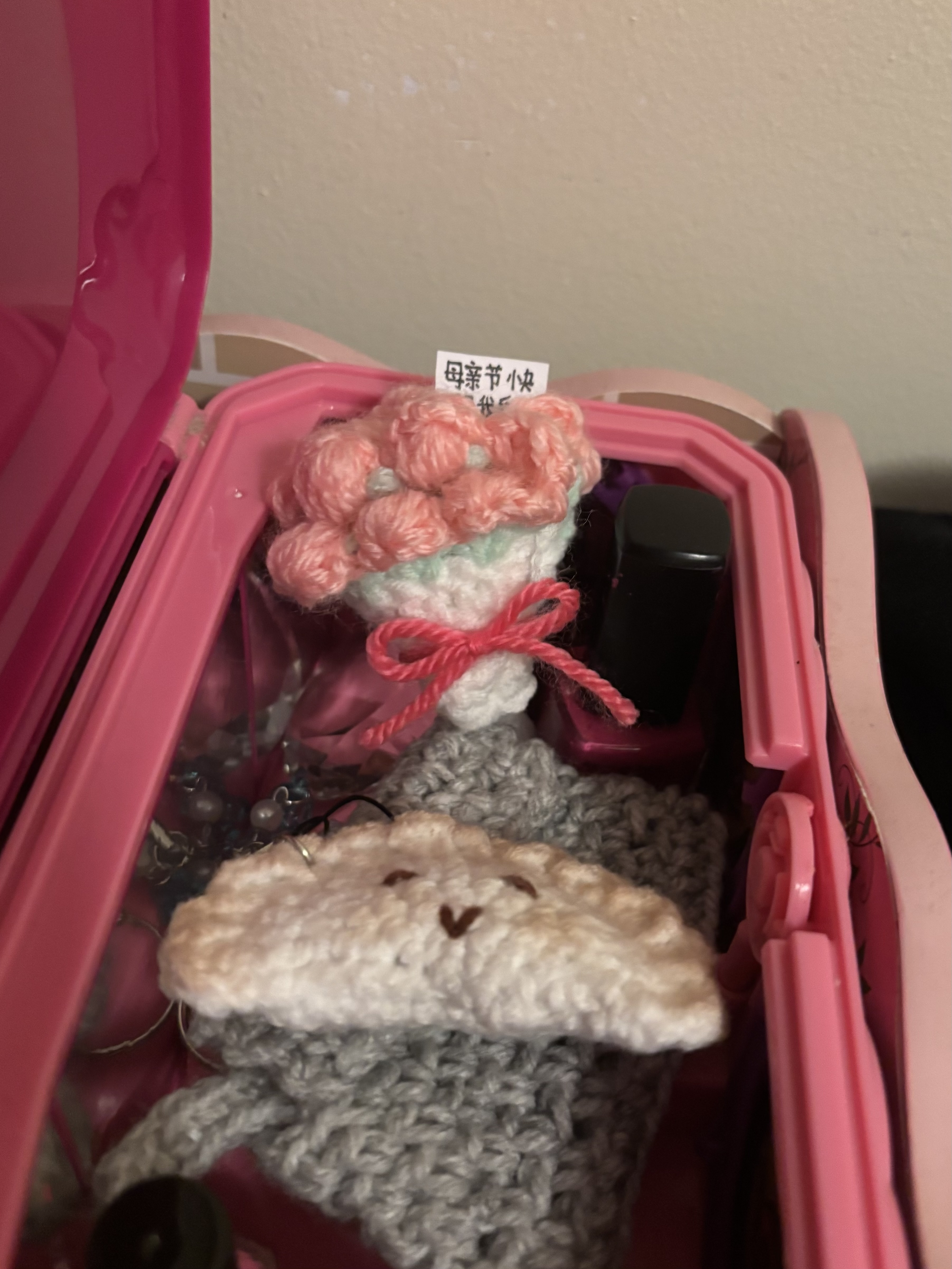 a closeup shot inside a pink container showcasing various crochet amigurumi items. there's a bouquet of pink flowers tied with a pink ribbon and a crochet dumpling with a cute smiling face. a bottle of nail polish is also visible. the items appear to be handmade gifts. a label with chinese characters is attached near the bouquet, possibly related to mother's day.