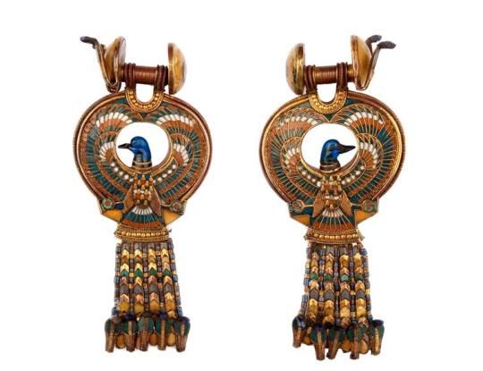 this image showcases a pair of ancient egyptian earrings. each earring features a falcon with outstretched wings, meticulously crafted from gold. the wings are adorned with vibrant inlays of lapis lazuli and turquoise. the body of the falcon is highlighted with colorful enamel, and a striking blue head adds to its majestic allure. the earrings dangle from intricate gold hoops, culminating in a cascade of finely crafted beads.